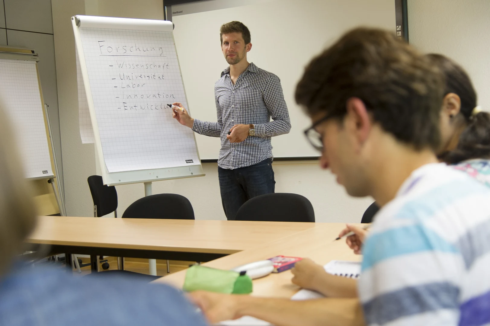 Eine Wissenschaftlerin und ein Wissenschaftler arbeiten hinter einer Glasfassade und mischen Chemikalien mit Großgeräten.