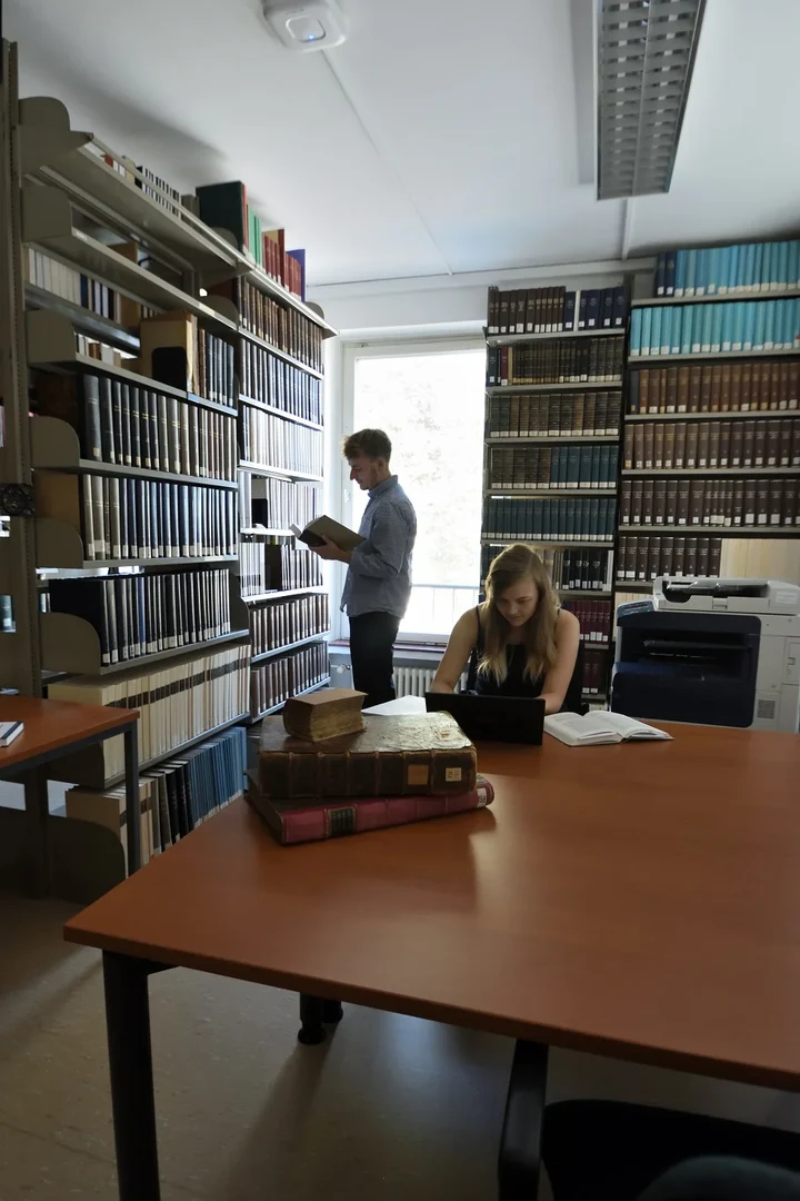 Eine Wissenschaftlerin und ein Wissenschaftler arbeiten hinter einer Glasfassade und mischen Chemikalien mit Großgeräten.