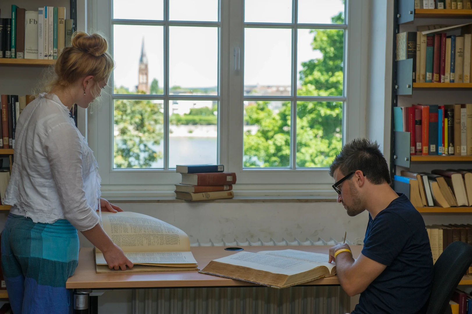 Eine Wissenschaftlerin und ein Wissenschaftler arbeiten hinter einer Glasfassade und mischen Chemikalien mit Großgeräten.